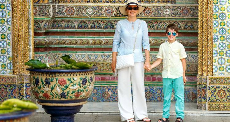 Mother and son pose happily in front of ornate, mosaic-covered temple walls in Bangkok wearing sun hats and sunglasses.