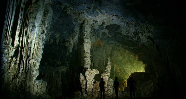 Caverna poco iluminada con estalactitas y estalagmitas gigantes iluminadas por focos que revelan texturas dramáticas.