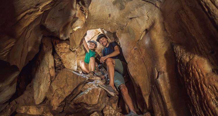 Dos aventureros sonríen mientras trepan por una estrecha abertura de roca dentro de una cueva, llevando cascos y arneses.