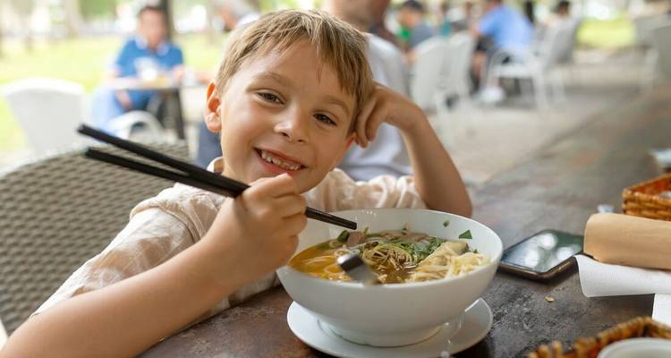 Un niño sonriente come un humeante tazón de pho vietnamita con palillos en una cafetería al aire libre.