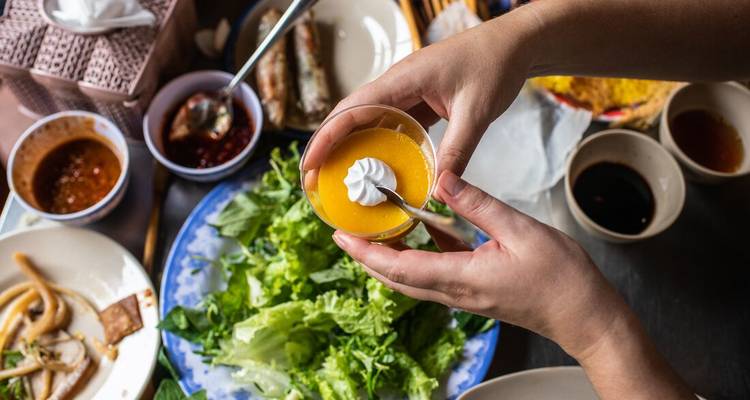 Primer plano de manos sosteniendo un pequeño postre sobre una colorida comida vietnamita con salsas y vegetales frescos