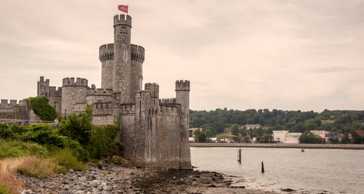 Medieval stone castle with circular tower and red flag overlooks a quiet estuary under cloudy sky.