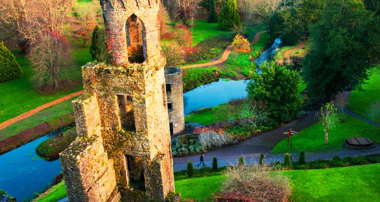 Ruins of a medieval stone tower rise above lush green parkland and winding stream in golden light