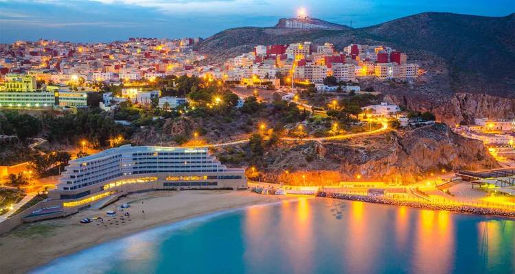 A vibrant coastal city at dusk with hillside houses and their lights reflecting in a calm bay.