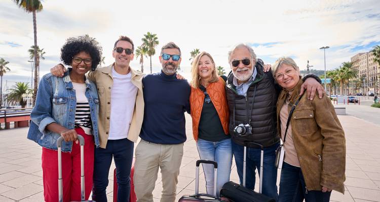 A diverse group of mature travelers in casual attire smile together with luggage along a sunny palm-lined promenade.