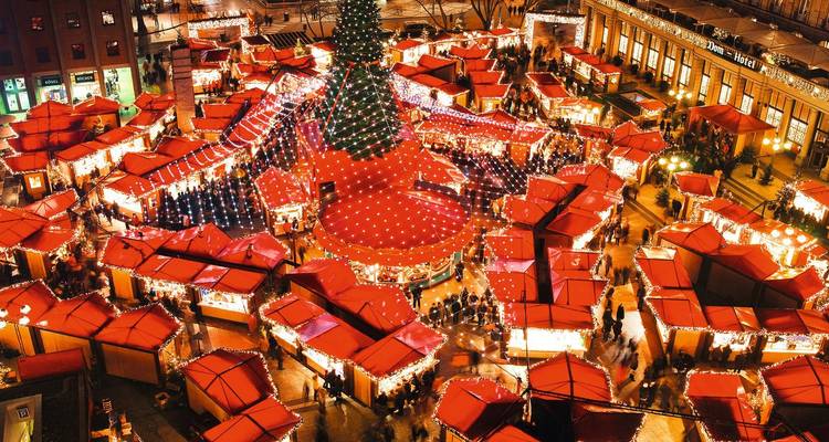 Vibrant aerial view of a European Christmas market with red-roofed stalls surrounding a giant tree.