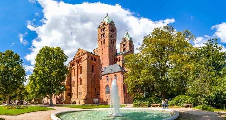 Romanesque red-brick cathedral with twin towers behind a round fountain and green park.