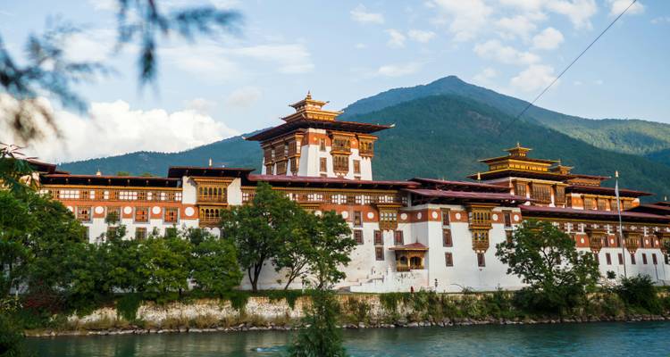Les murs blancs et rouges du Punakha Dzong s'élèvent au-dessus de la rivière turquoise avec des montagnes boisées en arrière-plan.