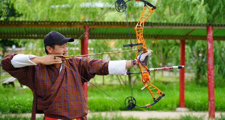 Un archer bhoutanais vêtu du gho traditionnel vise avec un arc de compétition orange vif dans un champ de tir extérieur.