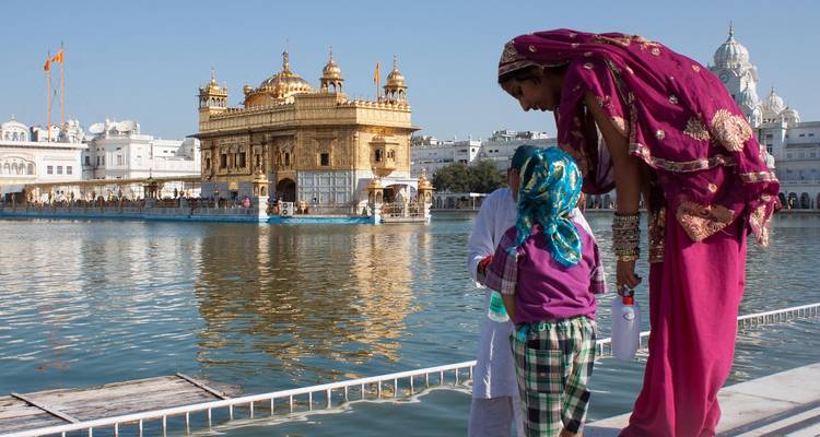 Woman in vibrant sari and child stand on marble edge overlooking the Golden Temple across calm water.