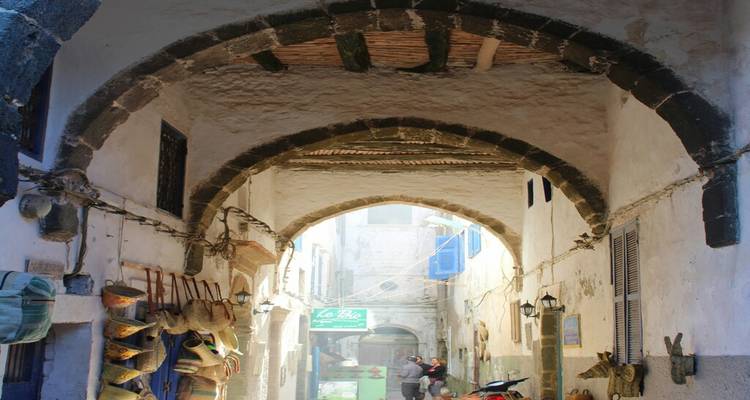 Les arcades en pierre ensoleillées encadrent une ruelle étroite de la médina bordée de paniers suspendus et d'artisanats locaux, tandis que deux personnes se promènent dans la lumière chargée de poussière.