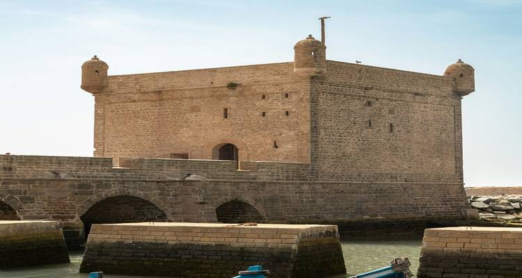 Une forteresse trapue et de forme carrée en pierre avec des tours d'angle s'élève au-dessus d'un pont en pierre et d'une eau de marée sous un ciel bleu dégagé.
