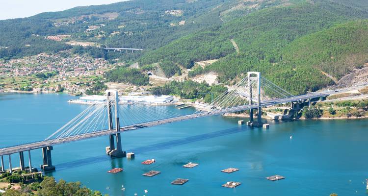 Cable-stayed Rande Bridge spanning a blue estuary surrounded by forested hills.