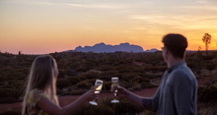 Couple en silhouette trinquant des verres de champagne au coucher du soleil avec les dômes de Kata Tjuta à l'horizon.