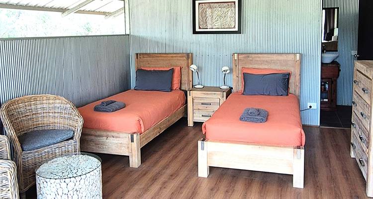 Chambre de cabane rustique avec deux lits jumeaux recouverts d'orange, sièges en osier et murs en métal ondulé.