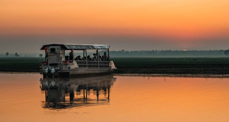 Un bateau de safari avec des touristes flotte sur une eau d'un calme miroir au lever du soleil avec un ciel orange.