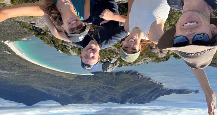 Group selfie atop a mountain overlooking the sweeping curve of Wineglass Bay and turquoise sea in Tasmania.