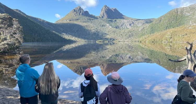 Five travellers stand on the shore of a still alpine lake admiring the dramatic twin peaks reflected in the water under a blue sky.