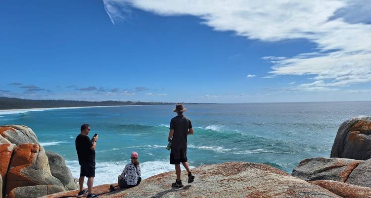 Three visitors stand and sit on orange-lichen granite boulders overlooking a turquoise sea beneath a wide blue sky.