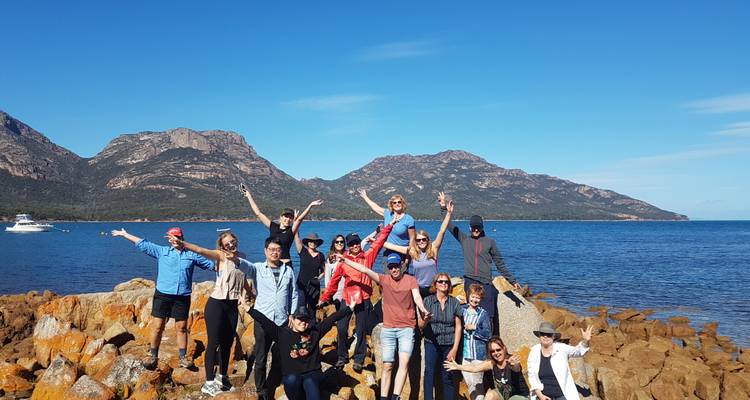 A large tour group enthusiastically poses on coastal rocks with blue sea and rugged mountains behind them.