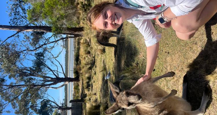 Smiling traveller kneels beside and gently pets a curious kangaroo in a grassy bush setting near a lagoon.
