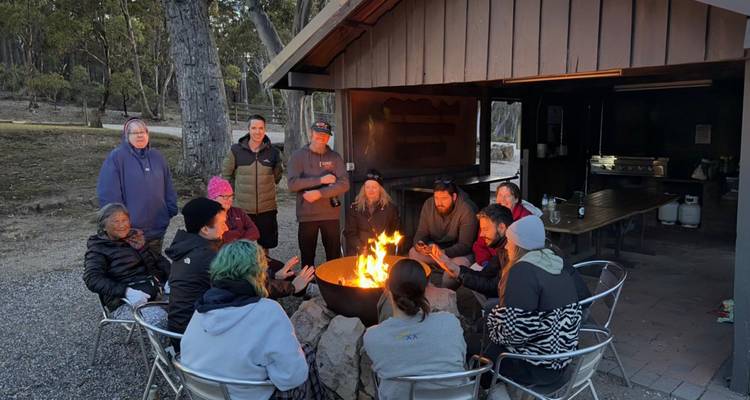 Travellers sit in a circle around a glowing campfire at an outdoor shelter in the evening.