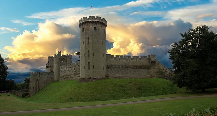 Warwick Castle’s massive round tower dominates grassy ramparts against a dramatic sunset sky with glowing clouds.