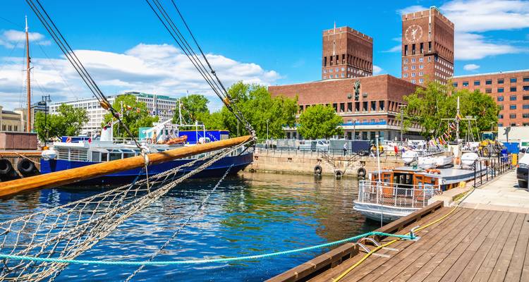 Sunny quay in Oslo with wooden sailing ships, blue water and the brick twin-towered City Hall behind.