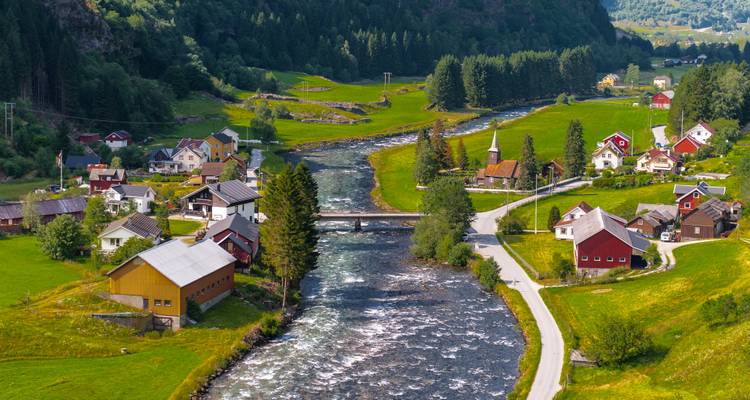 Idyllic Norwegian valley village with a river, bridge, white church and green meadows backed by forested hills.