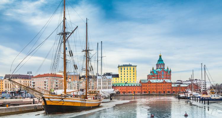 Historic tall ship moored in an icy harbour with colourful Helsinki waterfront buildings and Uspenski Cathedral behind.