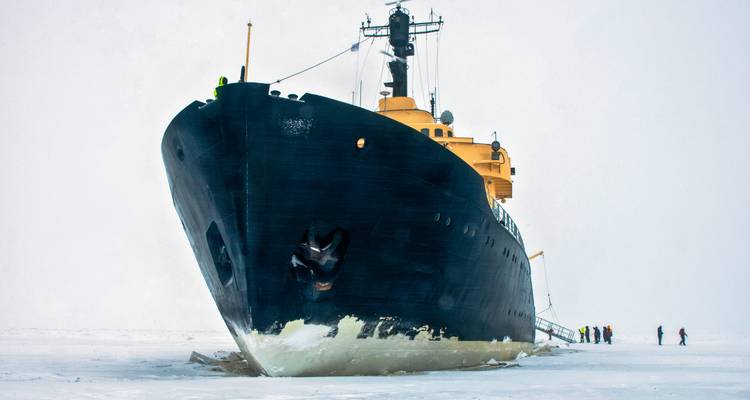 Massive icebreaker ship cutting through frozen sea with tiny figures walking beside the hull.