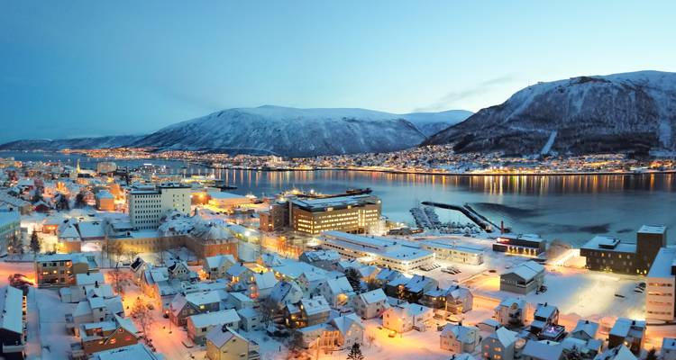 Tromsø cityscape at twilight with snow-covered rooftops and mountains reflected in calm harbour waters.