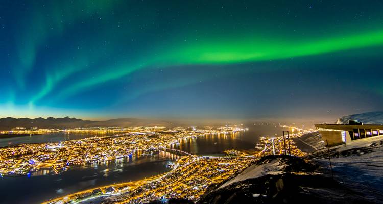 Brilliant green aurora sweeping above the night-lit city and fjord of Tromsø viewed from a snowy mountaintop.