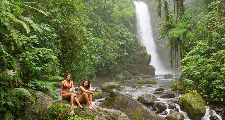 Two women in swimwear relax on rocks beside a lush jungle waterfall and river pool.