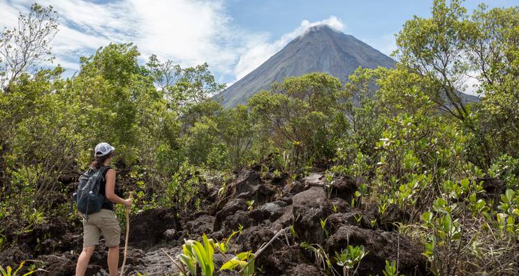 A hiker walks over black volcanic rocks toward the smoking cone of Arenal Volcano surrounded by lush forest.