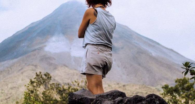A young woman stands on a rocky outcrop facing a dramatic conical volcano rising above rugged slopes and sparse vegetation.