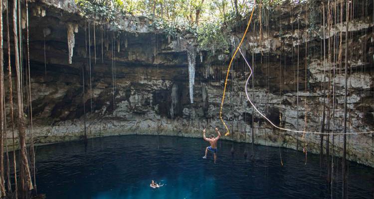 Adventurer swinging on a rope into the deep blue waters of a cavernous cenote lined with hanging vines.