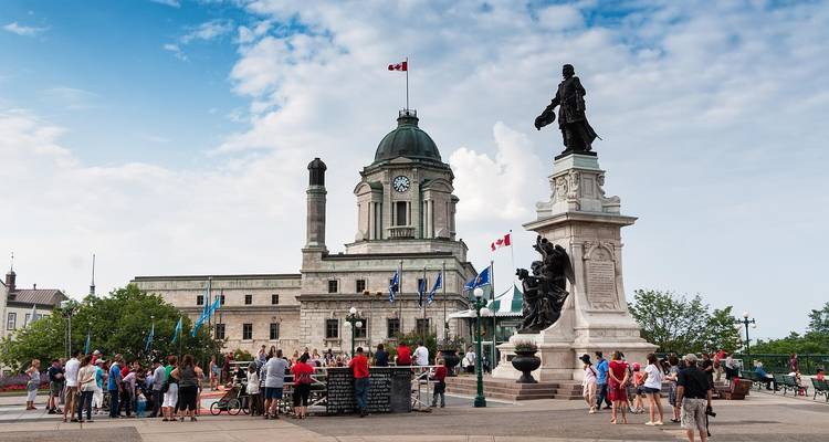 Des foules se rassemblent autour de statues sur une plaza devant un bâtiment patrimonial à dôme arborant des drapeaux canadiens.