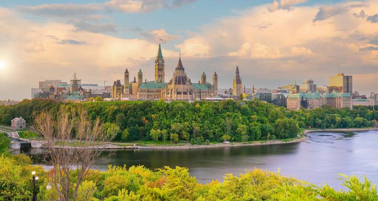 Les édifices du Parlement d'Ottawa s'élèvent au-dessus d'arbres d'un vert luxuriant lors d'un après-midi ensoleillé avec le fleuve au premier plan.
