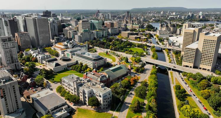 Vue aérienne diurne du centre-ville d'Ottawa présentant des immeubles modernes, des parcs verdoyants et le canal Rideau.