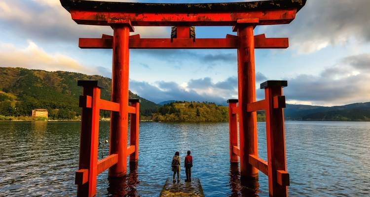 Vermilion torii gate of Hakone Shrine standing in Lake Ashi with two visitors admiring the view.