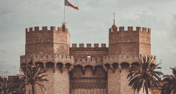 Die mittelalterlichen Steintürme Serranos in Valencia erheben sich über Palmen unter weichem Nachmittagslicht und einer wehenden Flagge.