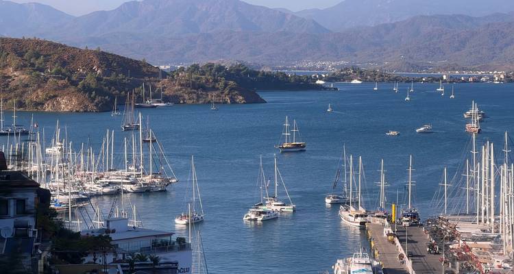 Vue sur le port avec des rangées de voiliers amarrés sur une eau bleu profond encadrée par des collines boisées et des montagnes lointaines.