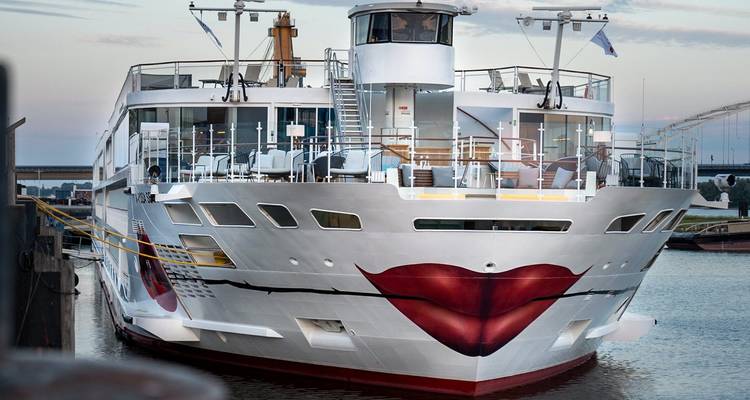 A modern river cruise ship with a stylised red lips design moored at a city quay.