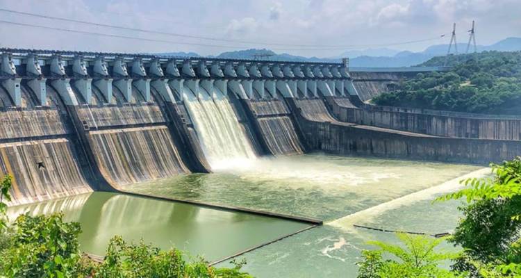 A large concrete gravity dam with water cascading over multiple spillways into a reservoir.