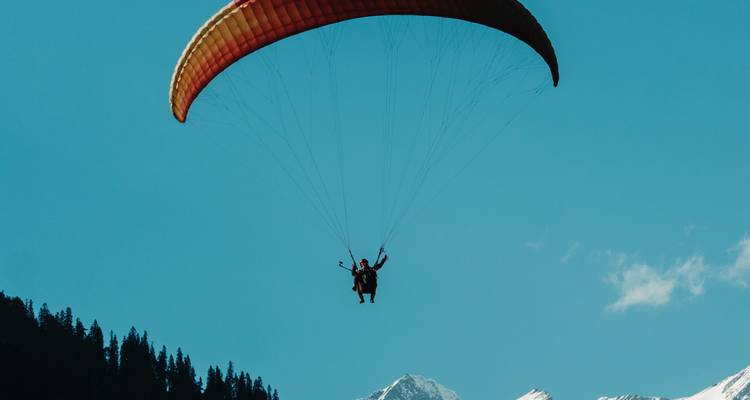 Un parapente en tandem s'élève haut dans un ciel bleu clair avec des montagnes enneigées en dessous.