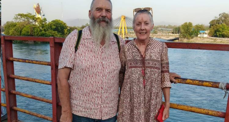 Un couple âgé sourit sur une passerelle au bord d'une rivière avec des manèges visibles au loin.