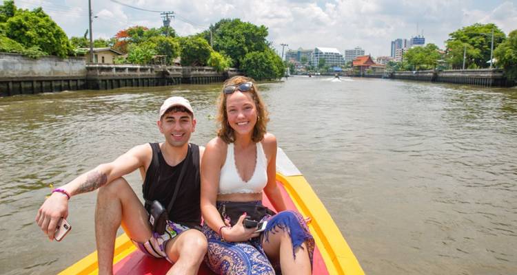 Una pareja joven sonríe en la proa de un bote de canal que navega por una vía fluvial tropical de la ciudad.