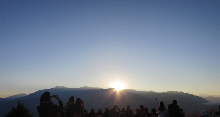 Menschenmenge in Silhouetten, die den Sonnenaufgang über entfernten Bergen unter klarem blauem Himmel beobachtet.