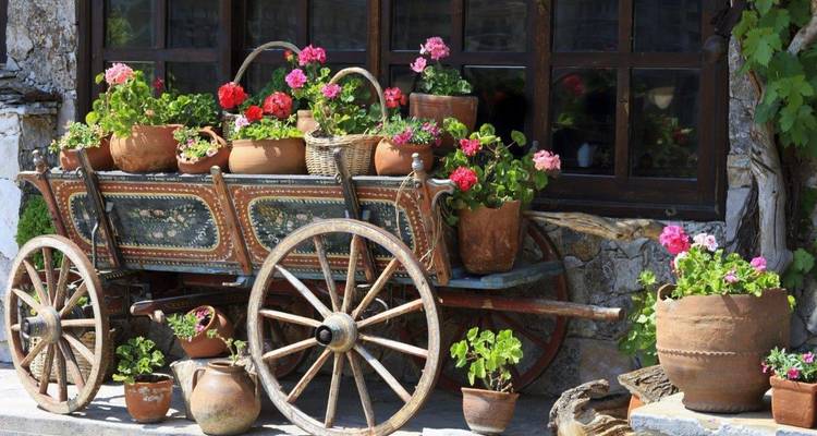 Chariot en bois décoratif débordant de pots en terre cuite remplis de fleurs aux couleurs vives devant une maison en pierre.